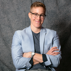 Ali Greey in a light blue-gray blazer jacket with a dark blue-gray shirt, in front of a dark gray background.