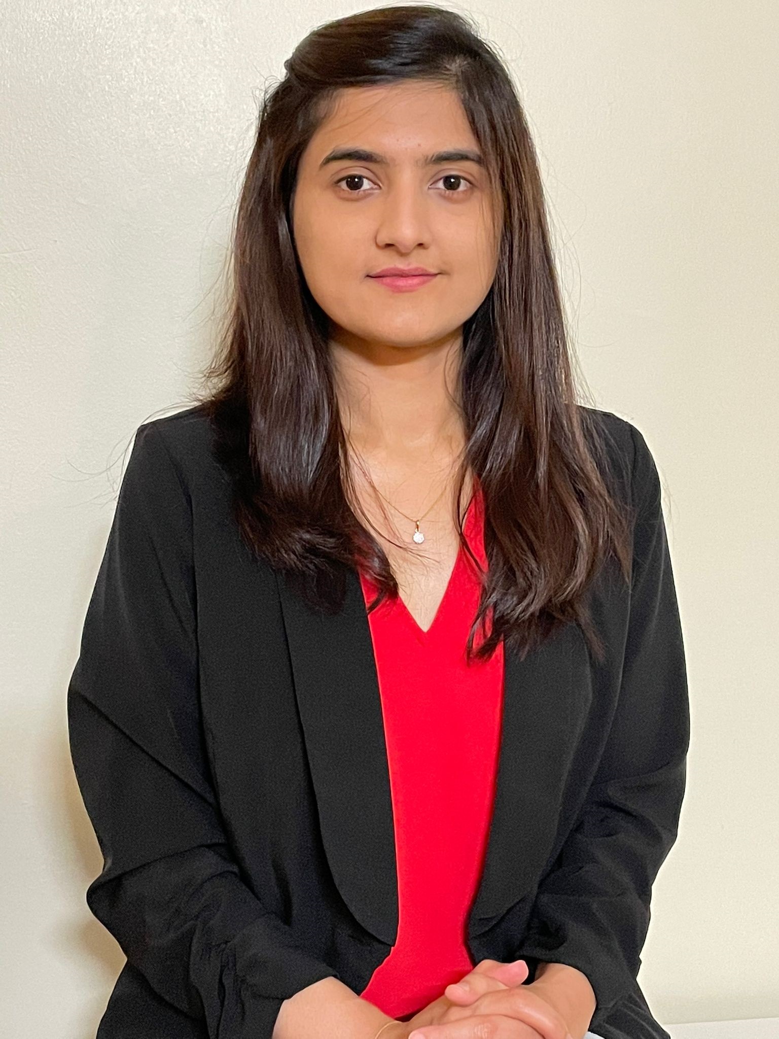 Anam Shahil Feroz in a red top under a black blazer, sitting in front of a cream-coloured wall.