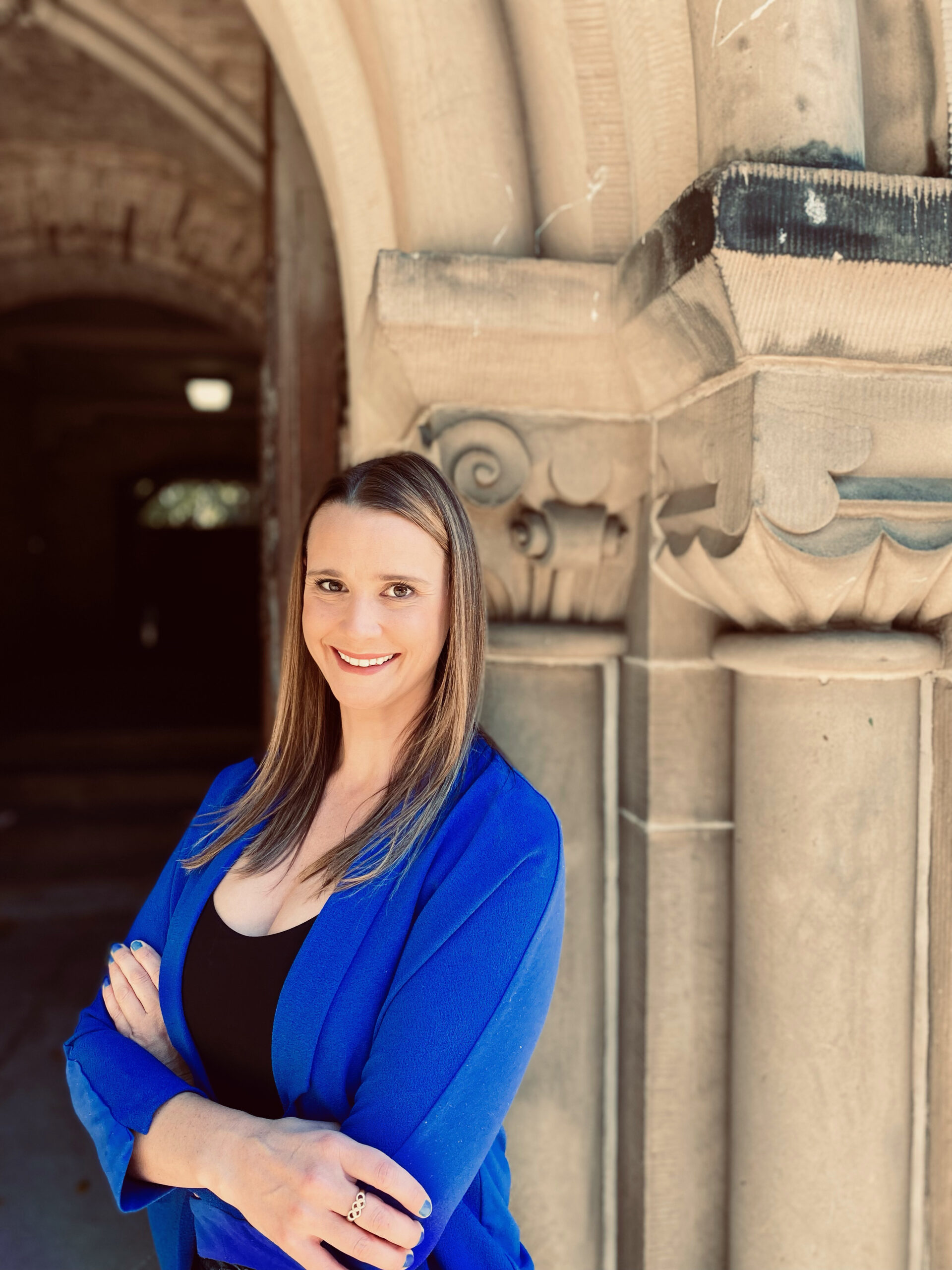 Erin Willson in a bright blue top, with a black top underneath. Erin is in front of a supporting pillar of an archway.