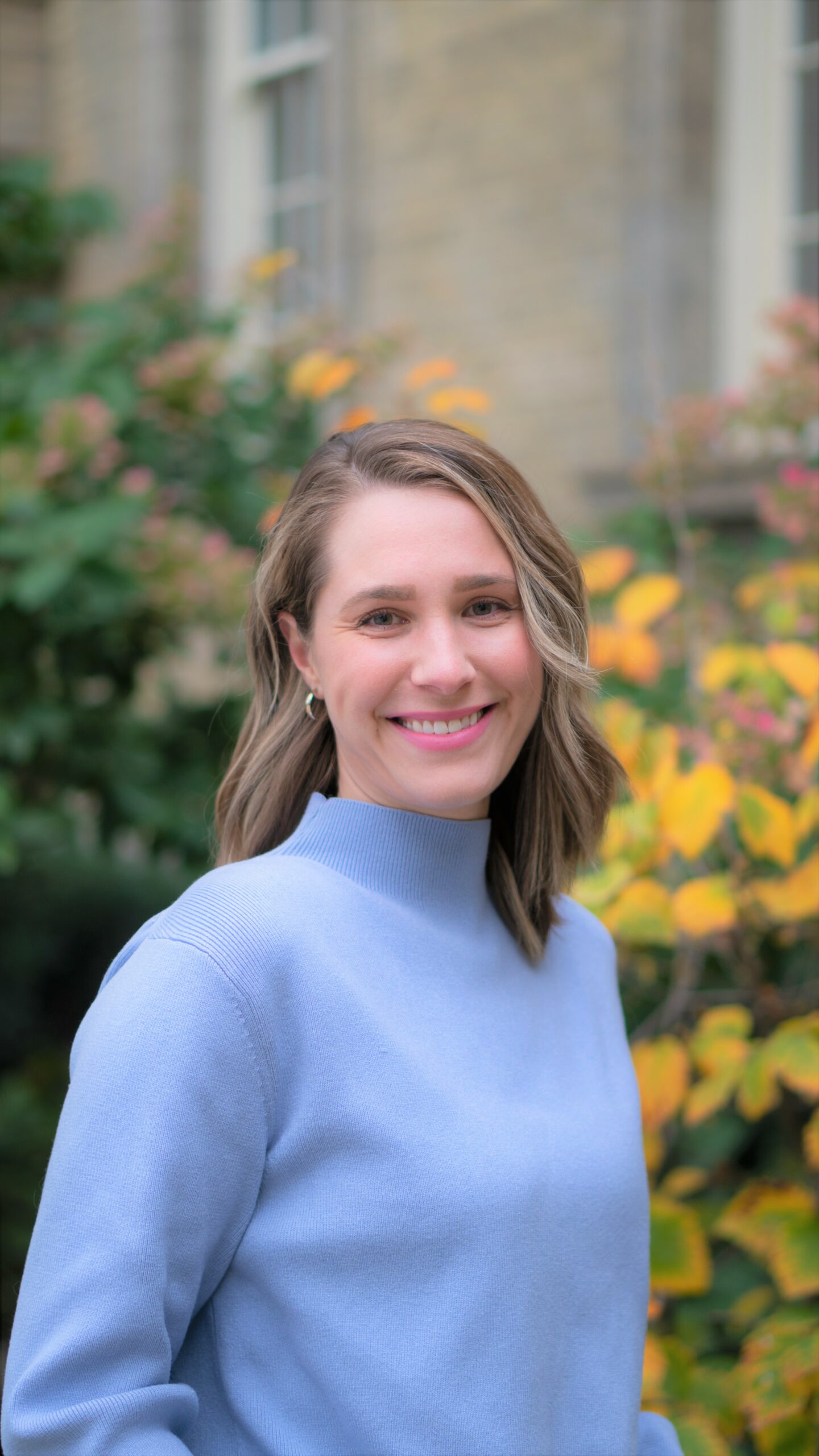 Madison Giles in a sky blue sweater in front of a blurred out background of green-yellow leaves in front of a building.