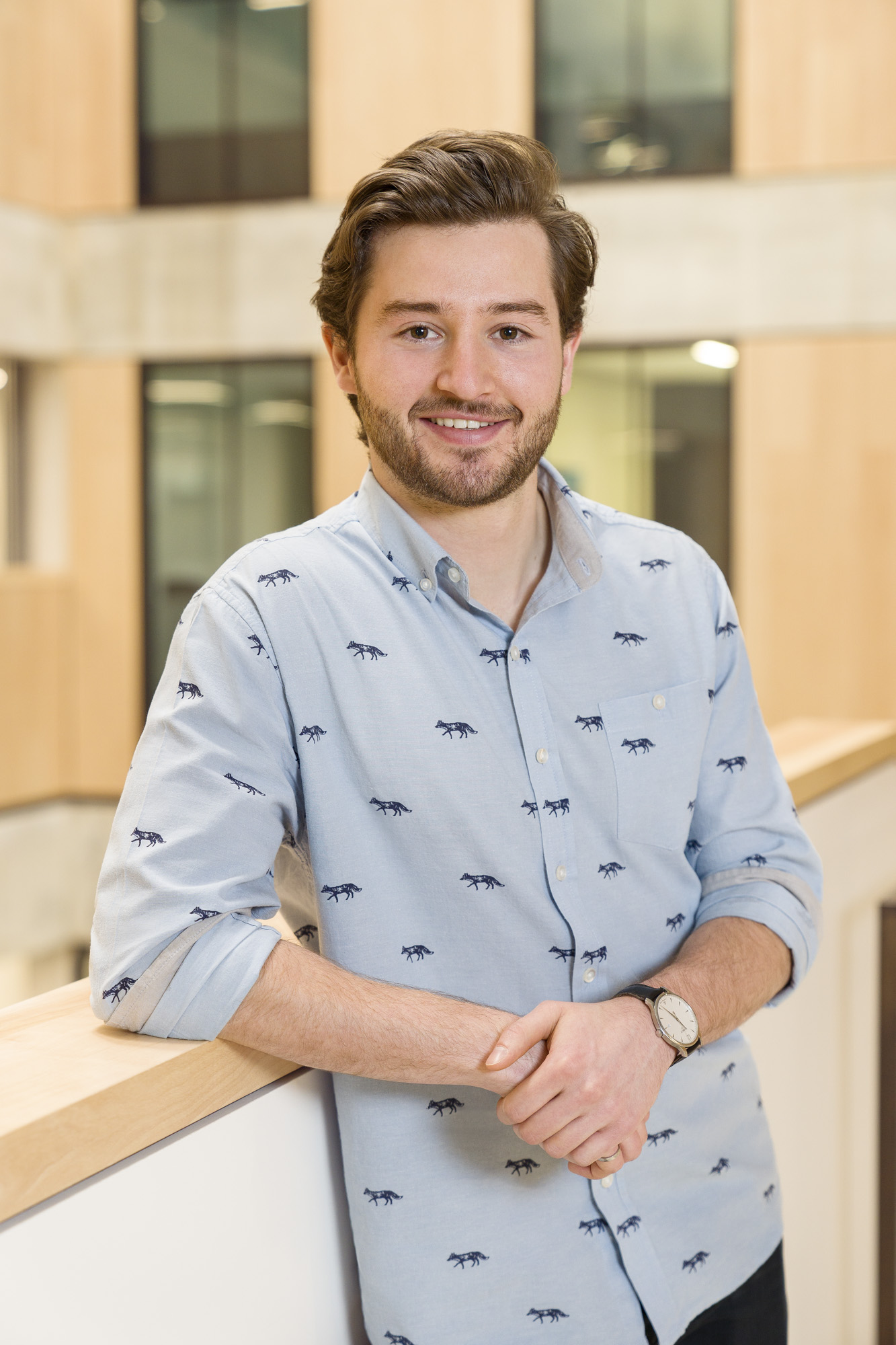 Peter Sterles wearing a light blue collared shirt with a pattern of foxes. Peter is leaning on a railing inside of a brightly-lit building.