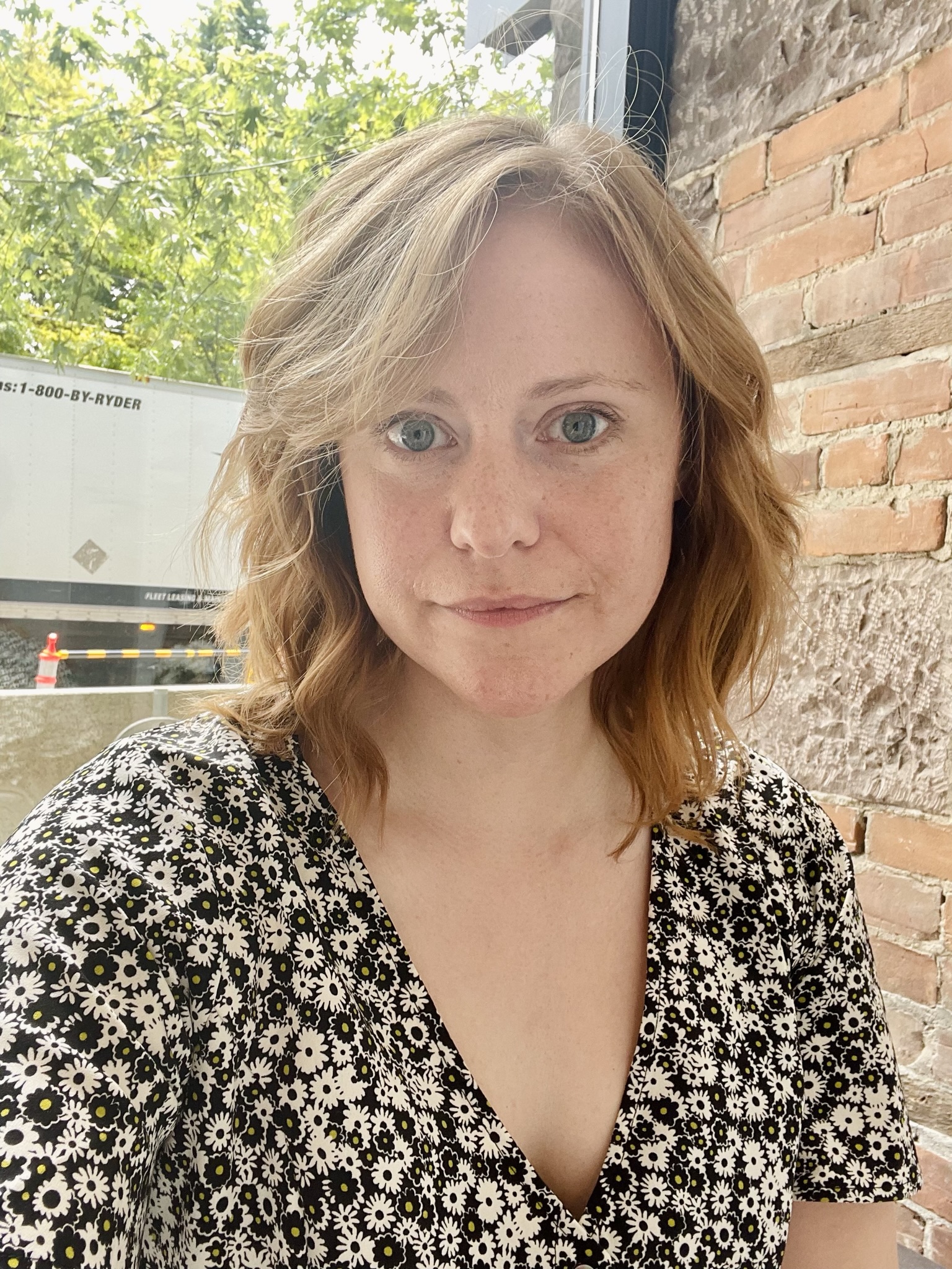 Rose Schmidt in a flower-patterned top, in front of a brick wall in the right foreground, and trees in the left background.